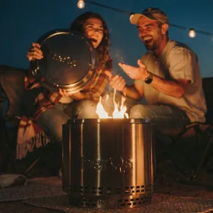 herQs Cozy fire pit burning outdoors, with a metal lid tray held beside two seated people