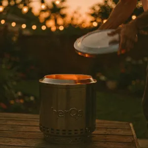 herQs Cozy Closer fire pit lid shown over a black cylindrical fire pit, with the lid held above it outdoors.