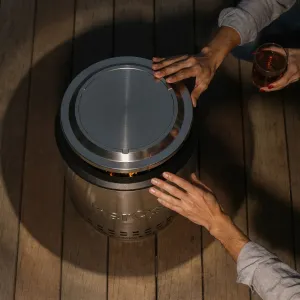 herQs Cozy fire pit lid on a round tabletop fire pit, with flame visible beneath the brushed metal cover