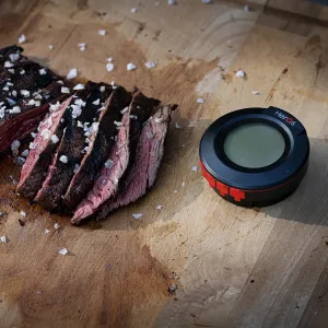 herQs Kuppelthermometer mit rundem Display und roten Tasten neben geschnittenem Grillfleisch auf einem Schneidebrett