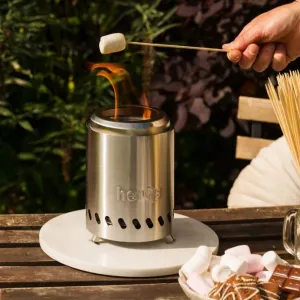 herQs smokeless tabletop fire pit burning on a wooden table with a marshmallow skewer and roasting snacks nearby.