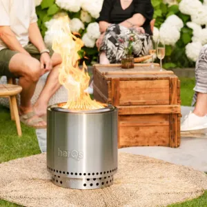 herQs rauchfreie Feuerstelle mit sichtbarer Flamme, Edelstahlgehäuse und Logo, im Freien für gesellige Runden aufgestellt