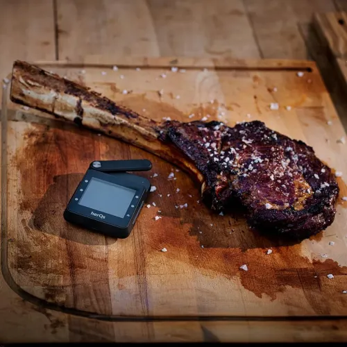 HerQs termometer, svart kvadratisk enhet med skärm, knappar och utfälld arm, ligger på skärbräda bredvid en stor tomahawk.