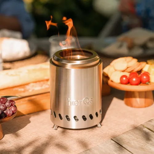 Kompakti, sylinterimäinen pöytätakka harjatusta teräksestä herQs:ltä, jossa on avoin yläosa, liekki, tuuletettu jalusta ja pienet jalat, puisen ulkopöydän keskellä.