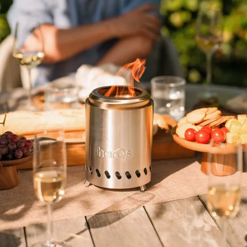 herQs Smokeless FirePit Mini ruostumattomasta teräksestä, jossa on pyöreä aukko, ilmanvaihtoaukot ja pienet jalat, asetetaan pöydälle.
