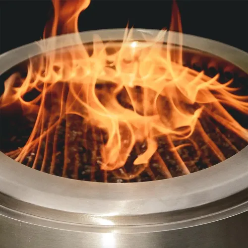 herQs Rökfri Eldstad Mighty i blankt stål med rund kant, synligt innergaller och höga lågor som brinner inuti öppningen.