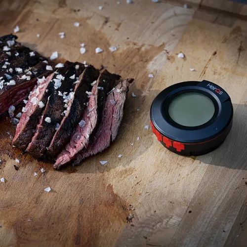 herQs Kuppelthermometer mit rundem Display und roten Tasten neben geschnittenem Grillfleisch auf einem Schneidebrett