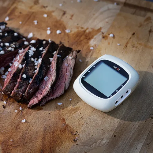 herQs EasyBBQ trådlös termometer med appnotiser bredvid skivat grillat kött på en träbräda