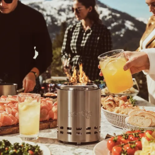 Cylindrisk eldstad i rostfritt stål med herQs-logotyp, övre öppning med låga och ventilationshål, på litet utomhusmatbord.