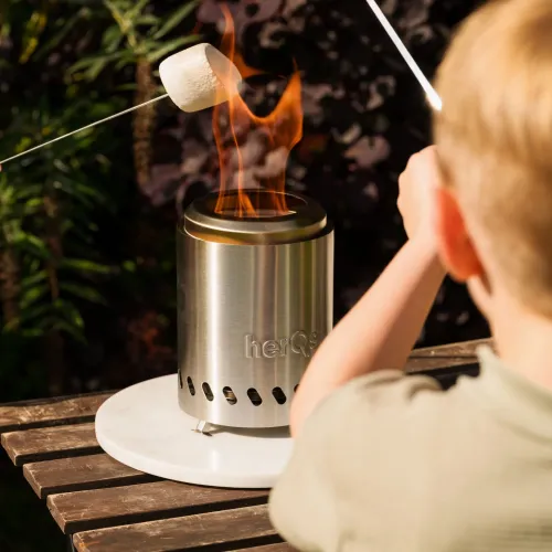 herQs FirePit Mini, lieriömäinen teräksinen tulipesä, jossa on liekit ja soikeat tuuletusaukot, pyöreällä tarjottimella ulkopöydällä; vaahtokarkki vartaassa.