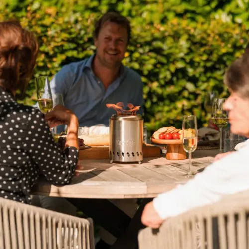 herQ:n savuton FirePit Mini, joka on ruostumattomasta teräksestä valmistettu lieriömäinen, rei'itetyillä tuuletusaukoilla ja näkyvällä liekillä, seisoo ulkopöydällä.