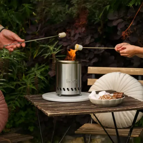 Kompakti savuton ruostumattomasta teräksestä valmistettu takka, jossa on herQs-logo; lieriön muotoinen, jossa on tuuletusreiät ja jalat, pöydällä, jossa on näkyvä liekki.