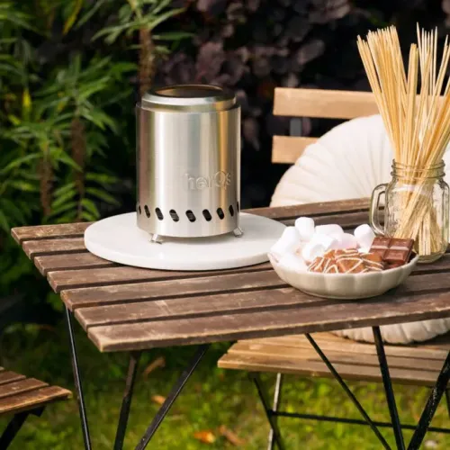 herQs Smokeless FirePit Mini harjatusta teräksestä, lieriömäinen, tuuletusrakoilla ja pienillä jaloilla, valkoisella levyllä puupöydällä.
