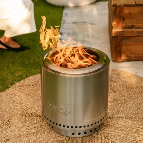 herQs FirePit Cozy in brushed stainless steel, cylindrical fireplace with top edge and flames, ventilation holes around the base outdoors.