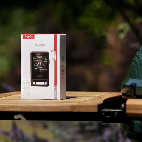 herQs Pin PRO Fleischthermometer-Verkaufsverpackung mit 2 kabellosen Sonden auf einem Gartentisch neben einem Grill